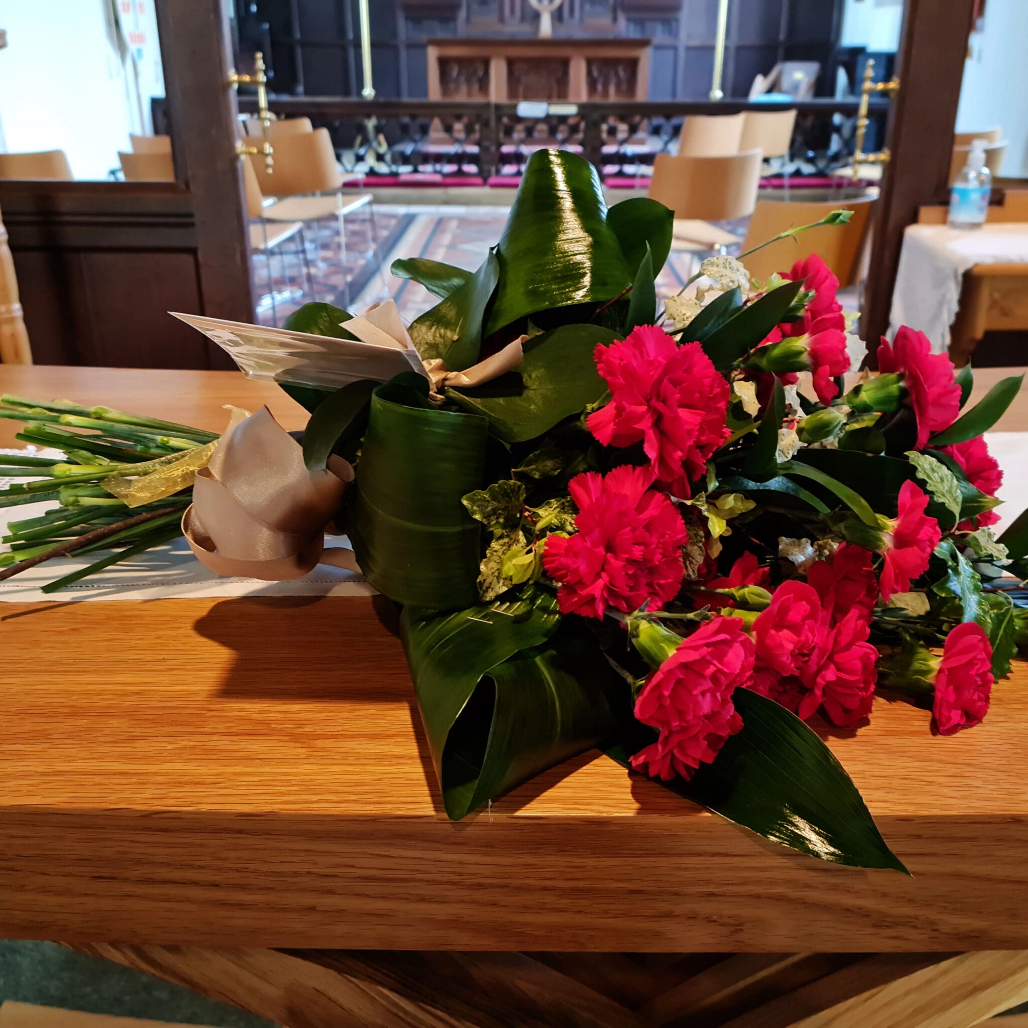 A bouquet of red flowers and green leaves rests on a wooden surface inside a church, with chairs, an altar, and other furniture visible in the background.