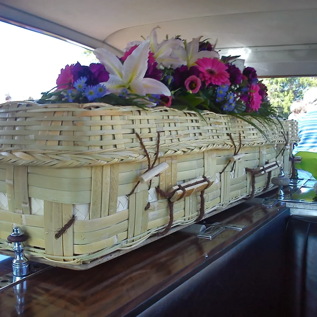 A woven wicker casket adorned with colorful flowers rests inside a vehicle, likely a hearse, suggesting a funeral or memorial service.